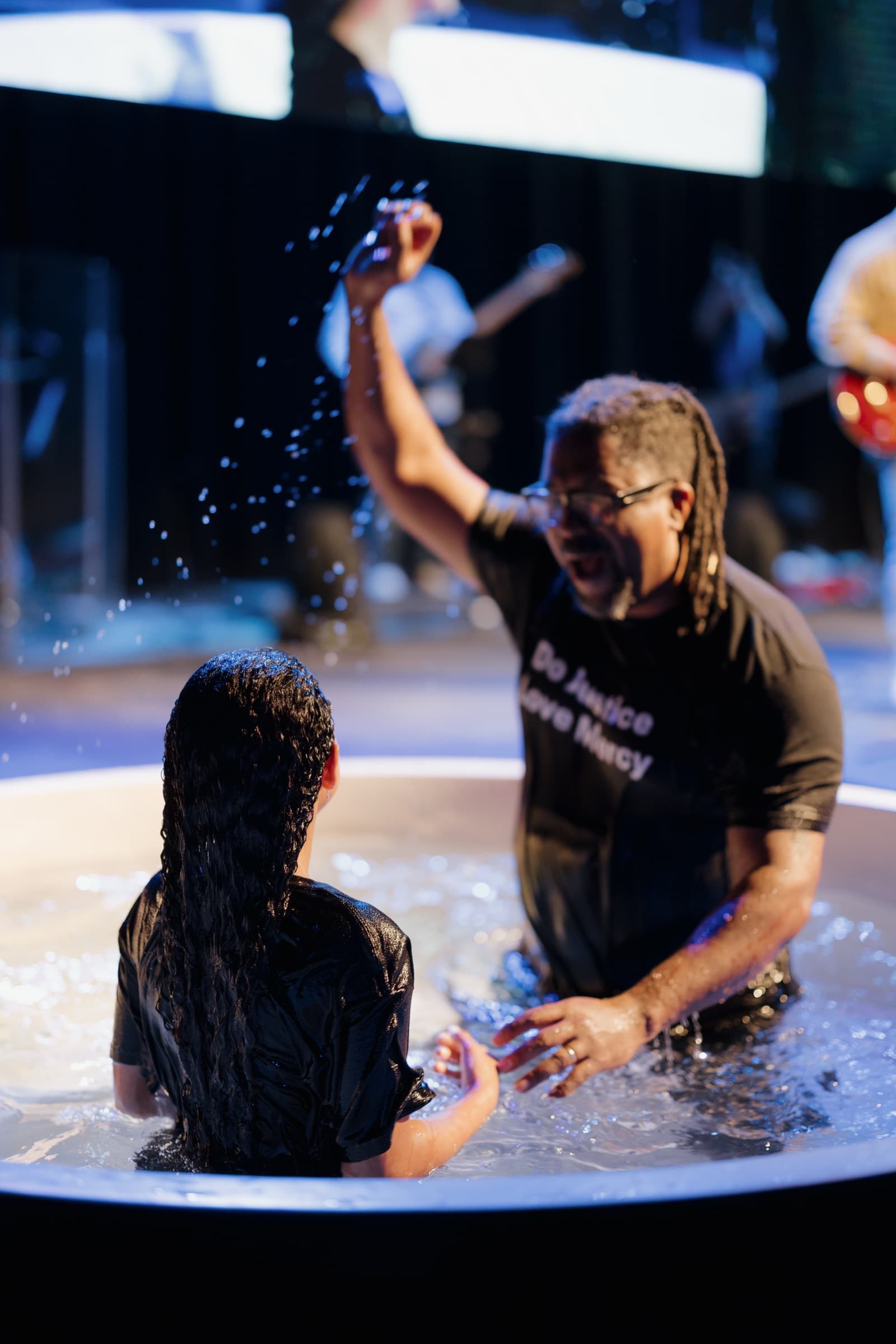 A pastor and a young woman celebrate in the baptism pool as water splashes.